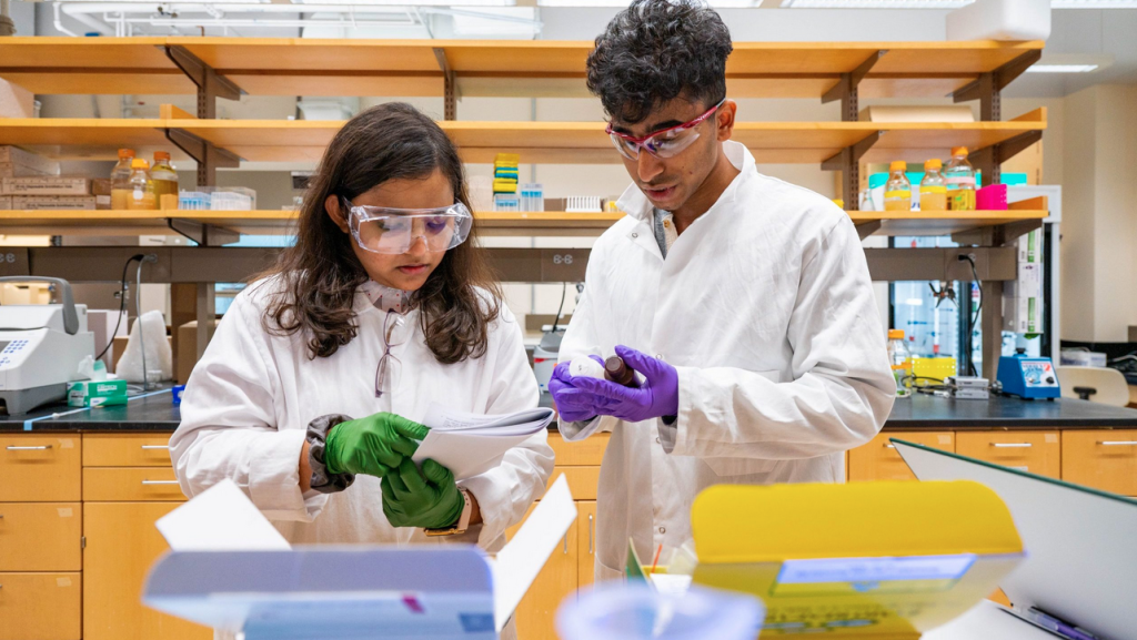 Two people in lab coats wearing safety googles hold chemicals and read a lab manual